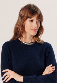Donkerblauwe gebreide trui met een ronde halslijn, vergezeld van een zilveren halsketting. Het model heeft schouderlang haar en gekruiste armen.