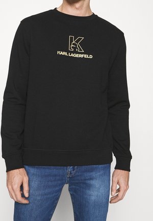 Sudadera negra con cuello redondo, mangas largas y un logo impreso en dorado, con una textura suave y puños y dobladillo acanalados.