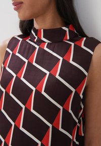 Korte mouw blouse in donkere stof met een geometrisch patroon van rode en witte driehoeken, hoge halslijn en een soepele textuur.