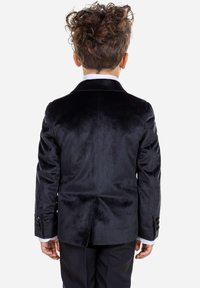 Blazer de terciopelo negro con una textura suave, con solapas de muesca y puños abotonados, llevado sobre una camisa blanca, visto desde la parte posterior.