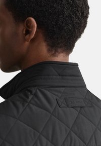 Zwarte doorgestikte jas met een hoge kraag, voorzien van ruitvormige stiksels en een gestructureerd oppervlak; logovignet op de achterkant nabij de kraag.