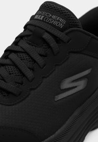 Μαύρο αθλητικό παπούτσι με αναπνεύσιμο επάνω μέρος από δίχτυ, υφή λεπτομερειών, επενδεδυμένο κολάρο και το λογότυπο "SKECHERS MAX CUSHION" στη γλώσσα.