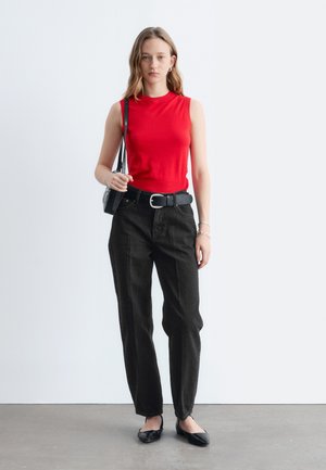 Kvinde iført en rød ærmeløs top, sorte højtaljede bukser med bælte, sorte flade sko, der holder en sort skuldertaske, stående på gråt gulv.