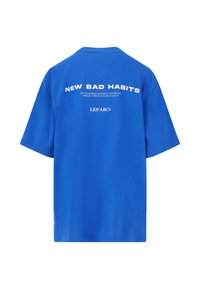 Sininen puuvillapaita rentoa kokoa, jossa on valkoista tekstiä selässä, joka lukee "NEW BAD HABITS" ja "LES'ARCS." Lyhyet hihat.
