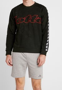 Uomo che indossa un felpa nera a maniche lunghe con il logo rosso "Lotto" e maniche a strisce nere, abbinata a pantaloni corti grigi chiari con il logo "First".