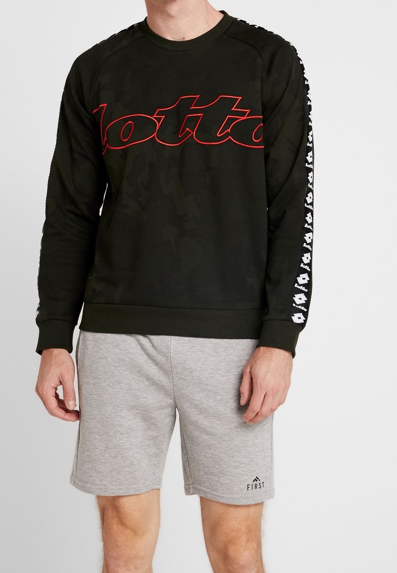Uomo che indossa un felpa nera a maniche lunghe con il logo rosso "Lotto" e maniche a strisce nere, abbinata a pantaloni corti grigi chiari con il logo "First".