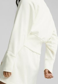 Felpa color crema chiaro con design cropped, polsini a costine e vestibilità rilassata, abbinata a pantaloni corti coordinati con un logo decorativo.