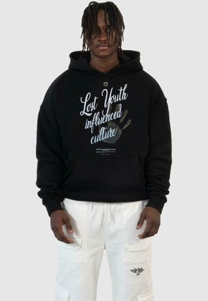 Sort hættetrøje med hvid og grå tekst, der står "Lost Youth påvirkede kultur," med et grafik af en hånd. Bæres med hvide cargo shorts.