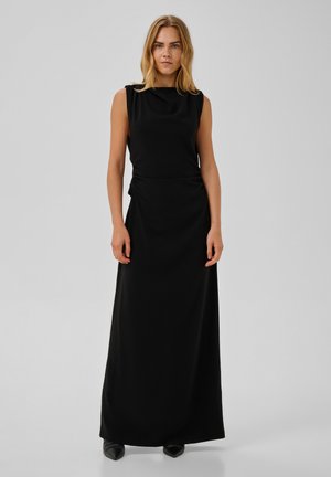 Femme aux cheveux blonds portant une robe noire sans manches longue jusqu'au sol et des chaussures noires pointues, debout devant un fond clair uni.