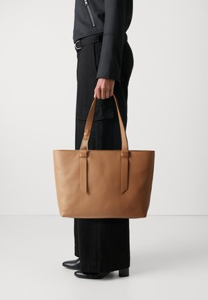 Skórzana torba tote w kolorze brązowym z teksturowanym wykończeniem, z dwoma paskami na ramię, prostokątnym kształcie i minimalistycznym designie. Noszona z czarnymi spodniami o szerokich nogawkach.