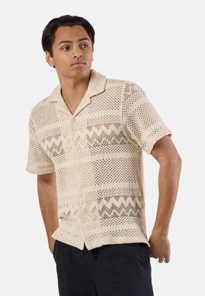 Jonge man met kort zwart haar draagt een beige gehaakt shirt met korte mouwen en zwarte broek, staand met zijn handen achter zijn rug.