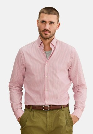 Man with short hair and light beard wearing a pink striped button-up shirt, gray undershirt, brown belt, and olive green pants, hands in pockets.