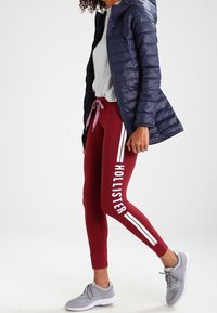 Leggings burdeos con franjas laterales blancas y texto, combinados con una camiseta gris, chaqueta acolchada azul marino y zapatillas deportivas grises.