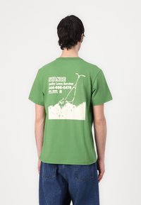 T-shirt de algodão verde com uma ilustração de um corta-relva nas costas, com texto branco para "Serviço de Cortar Relva Sortudo" e detalhes de contacto.