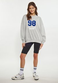 Szürke kapucnis pulóver kék "98" nyomattal, laza szabás, fekete kerékpáros short, fehér és fekete sportcipő, valamint csíkos zokni. Puha anyag, laza dizájn.