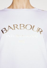Osoba v bílém tričku se lesklým zlatým nápisem "BARBOUR INTERNATIONAL" na hrudi a knoflíkovým detailem na rameni.