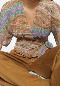 Trykt indpakningsbluse i let materiale med paisley- og stribemønstre i pastelfarver; parret med brune brede bukser.