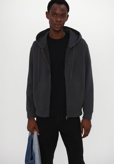 Gray zip-up hoodie with a drawstring hood, two front pockets, and ribbed cuffs. Worn over a black shirt and paired with dark pants.