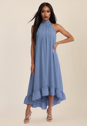 Frau trägt ein ärmelloses blaues Kleid mit hohem Kragen, gerafftem Saum und silbernen, mit Riemchen versehenen High Heels, steht mit einer Hand in der Hüfte.