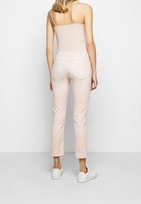 Jean skinny rose clair avec une texture lisse, taille mi-haute, deux poches arrière et jambes courtes, associé à des baskets blanches sur un fond neutre.