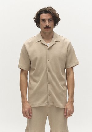 Mann mit lockigem Haar und Schnurrbart, der ein beige-farbenes, kurzärmeliges Hemd mit Knöpfen und passende Shorts vor einem einfarbigen, hellen Hintergrund trägt.