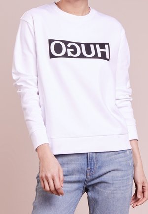 Person wearing a white long-sleeve sweatshirt with a reversed black "HUGO" logo and light blue jeans, standing against a plain background.