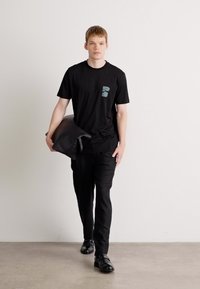 Schwarzes Baumwoll-T-Shirt mit einem tealfarbenen Taschenlogo, kombiniert mit schwarzen Hosen; hält ein graues Kissen, vor einem hellen Hintergrund.