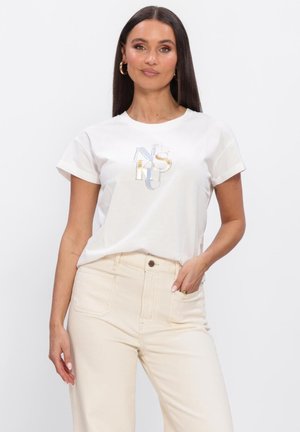 Vrouw met lang donker haar, goudkleurige oorringen, een wit T-shirt met korte mouwen en multicolor letters, en crème hoge taillebroek.