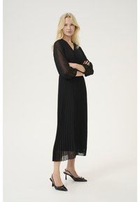 Schwarzes, plisseeartiges Kleid mit V-Ausschnitt, langen transparenten Ärmeln und Knöchellänge. Getragen mit schwarzen Slingback-Schuhen mit kleinen Verzierungen.