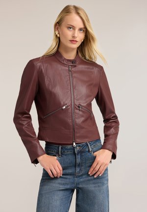 Frau mit langen blonden Haaren trägt eine braune Lederjacke mit vorderen Reißverschlüssen und blaue Jeans vor einem schlichten beigen Hintergrund.