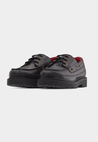 Μαύρα δερμάτινα παπούτσια με κορδό νια, με χοντρές λαστιχένιες σόλες, καφέ ραφές και κόκκινη εσωτερική επένδυση, εμφανίζονται σε ελαφριά γωνία πάνω σε λευκό φόντο.