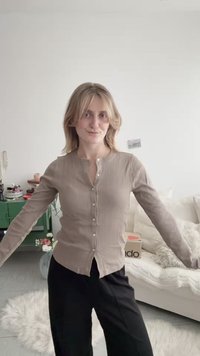 Νεαρή γυναίκα με ξανθά μαλλιά μήκους μέχρι τους ώμους φοράει μία μπεζ ζακέτα με κουμπιά και μαύρο παντελόνι, στέκεται με τα χέρια ελαφρώς ανοιχτά σε εσωτερικό χώρο.