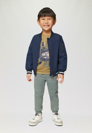 Marine bomberjack, beige graphic T-shirt, lichtgroene broek en witte sneakers. Heeft ritszakken en ribbels aan de mouwen.