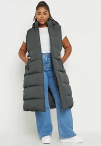 Chaleco acolchado gris con capucha, que cuenta con un cierre de cremallera de longitud completa, bolsillos laterales y un diseño esponjoso; llevado sobre una camiseta blanca y unos jeans azules.