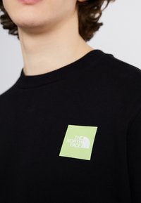 Primer plano de una persona que lleva una camiseta de cuello redondo negra con un logotipo cuadrado verde que dice "The North Face" en el pecho.