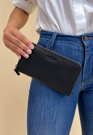 Black leather wallet with textured surface, zip closure, and embossed logo. Held against a backdrop of blue jeans and a white shirt.