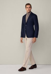 Blazer navy con due bottoni, abbinato a un dolcevita beige e pantaloni chiari. Mocassini marroni completano l'outfit, che mette in risalto un taglio classico.