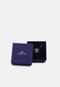 Collana Swarovski in una scatola viola, con un pendente circolare contenente una pietra nera circondata da cristalli trasparenti e una catena argentata.