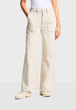 Žena oblečená v krémových širokých nohaviciach s veľkými prednými vreckami a béžovými šnurovacími čižmami, stojaca s jednou rukou vrecku.