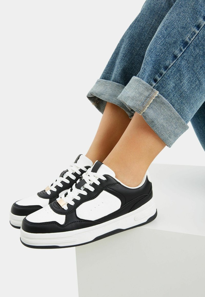 Black and white sneakers with leather and synthetic materials, featuring lacing and a textured rubber sole. Rolled denim jeans visible above.