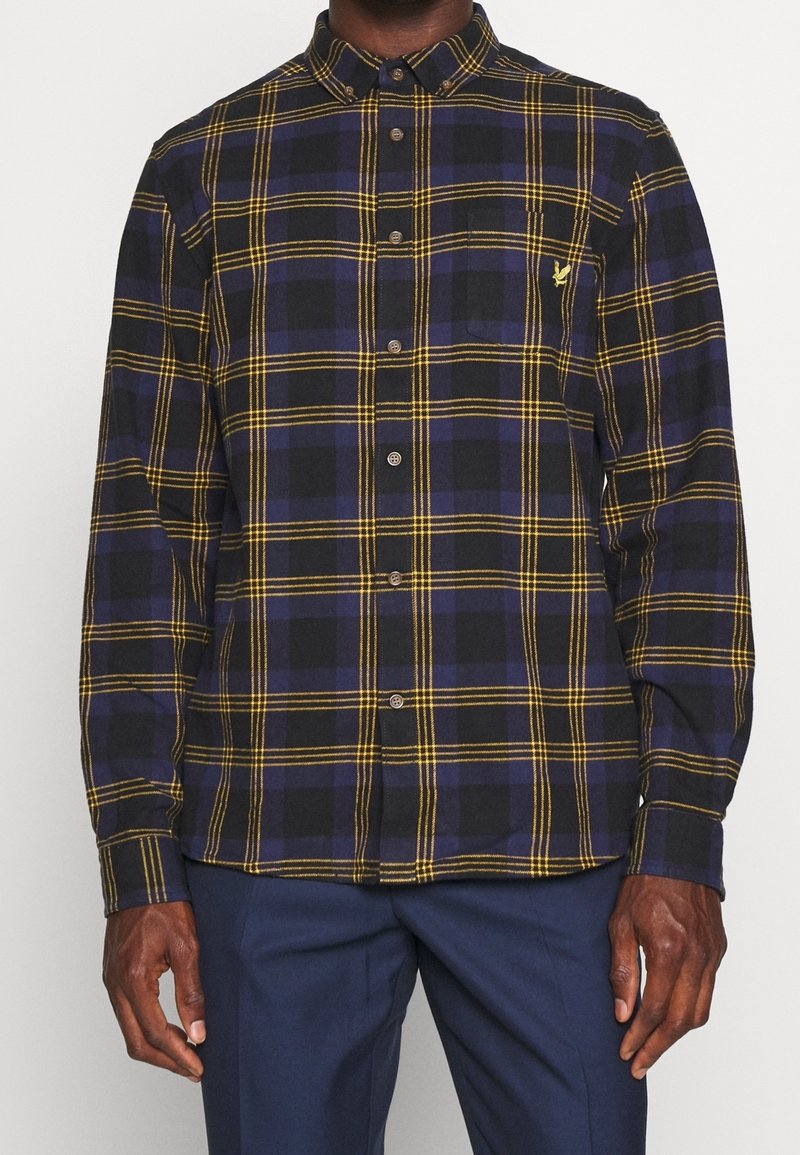 Chemise à carreaux bleu foncé et noir avec des lignes jaunes, boutonnée, portée avec un pantalon bleu marine, vue du cou jusqu'aux hanches.