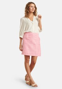 Witte blouse met subtiele gouden stippen, gecombineerd met een roze gestructureerde minirok met voorzakken. Beige enkelbandhakjes maken de outfit af.