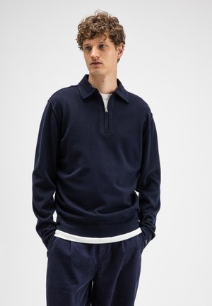 Man met krullend haar die een marineblauwe trui met rits draagt over een wit overhemd en marineblauwe broek, staande voor een effen lichte achtergrond.