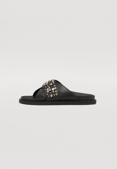 Schwarze Leder-Sandalen mit überkreuzten Riemen, verziert mit silbernen und goldenen Nieten auf dem Obermaterial, mit einer flachen Sohle und glatter Oberfläche.