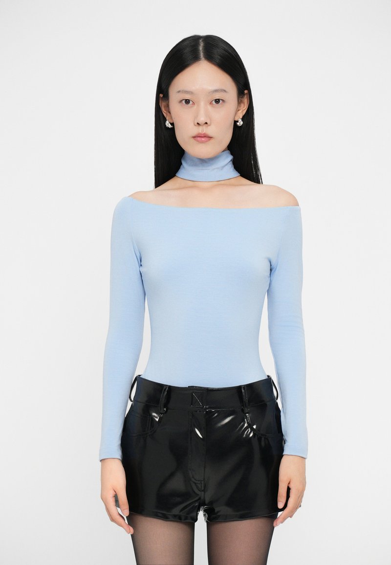 Světlomodrý off-shoulder body s dlouhým rukávem zkombinovaný s lesklými černými šortkami, které mají přiléhavý střih a hladkou texturu.