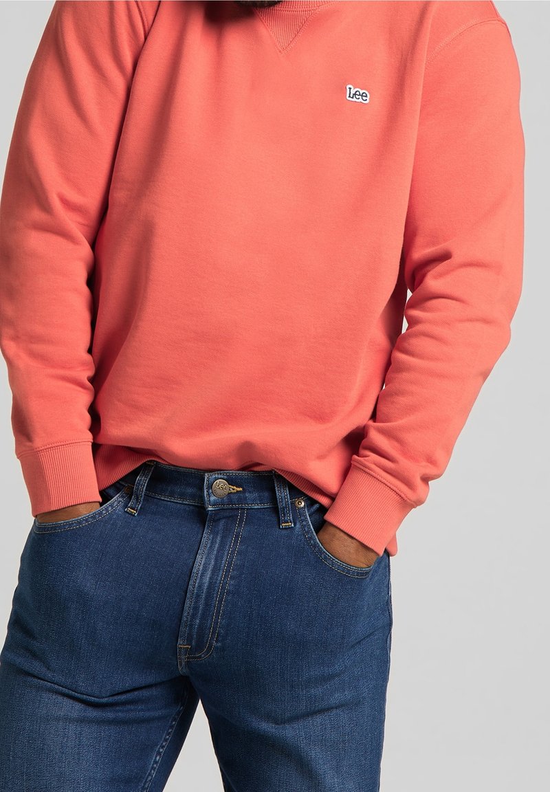 Koraal sweatshirt met ribbels aan de manchetten en de halslijn, voorzien van een klein blauw logo. Gecombineerd met donkerblauwe spijkerbroeken met minimale stiksels.