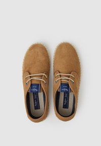 Zapatos de ante marrón con punta redonda, cordones de yute beige y una suela de esparto texturizada. El interior cuenta con un forro de tela oscura y una etiqueta.