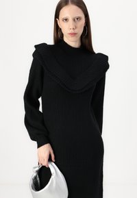 Juoda raštuota megzta suknelė su plačiu, volaniniu apykakle ir ilgomis rankovėmis. Dešinėje rankoje laikoma blizgi sidabrinė rankinė.