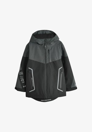 Jeune garçon aux cheveux bouclés portant une veste à capuche à carreaux noire et grise et un pantalon cargo de couleur claire, debout devant un fond uni.