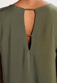Top vert olive avec un détail en trou de serrure dans le dos, fabriqué en tissu lisse. L'encolure présente une subtile forme en V. Design minimaliste avec des lignes épurées.